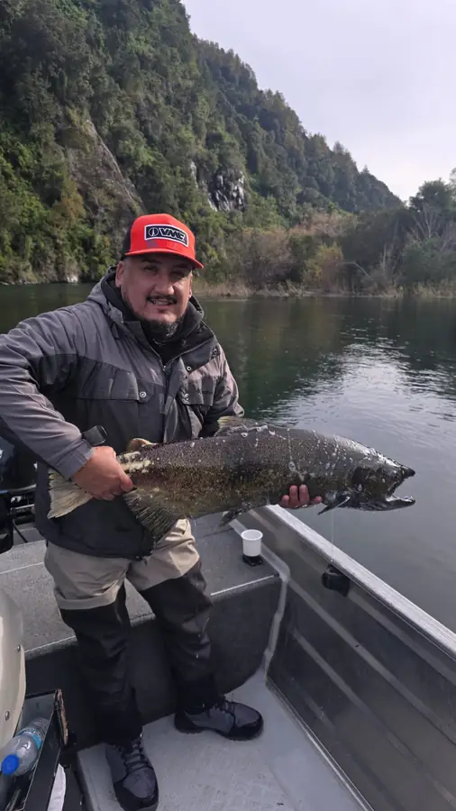 Socio del club mostrando una trucha grande, con la ladera del lago al fondo