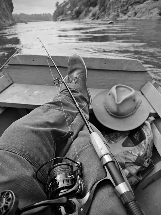 Caña y carrete apoyados sobre las piernas de un pescador en el bote, fotografía en blanco y negro